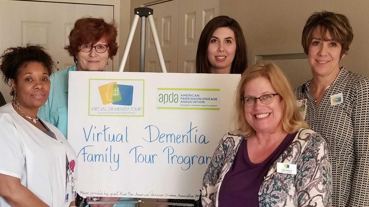 Staff pose for a photo in front of a sign that reads Virtual Dementia Family Tour Program