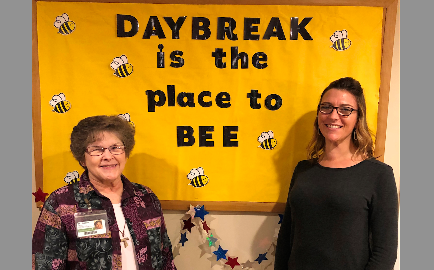 Two women pose in front of a photo that says Daybreak is the place to BEE